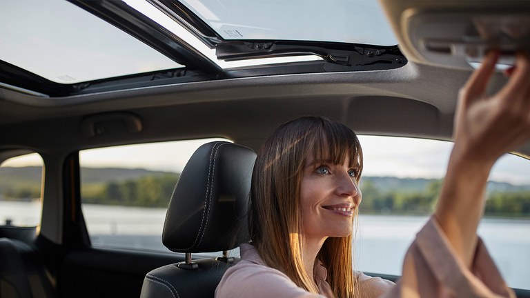 Frau mit langen braunen Haaren nun die Funktion des Panorama-Glasschiebehubdachs im Suzuki Vitara Hybrid.