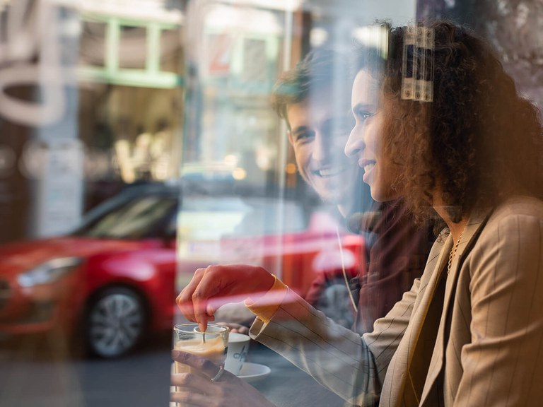 Ein Mann und eine Frau sitzen im Cafe am Fenster, im Hintergrund steht ein Suzuki Swift Hybrid in Burning Red Pearl Metallic.