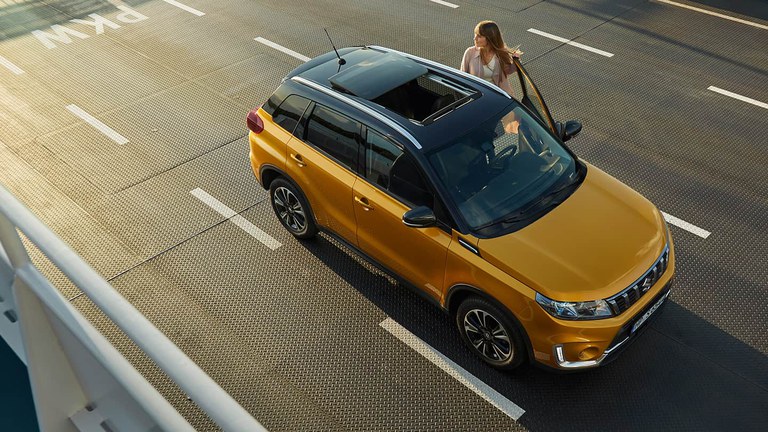 Ein Suzuki Vitara Hybrid in Solar Yellow Pearl Metallic auf einer leeren Fähre, von oben fotografiert. Eine Frau steigt ein.