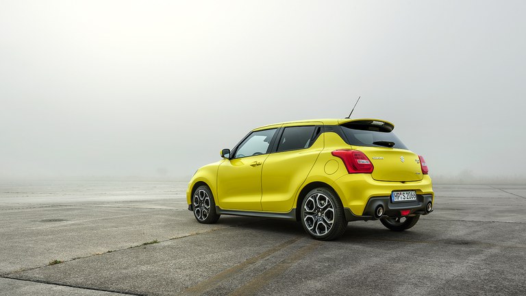 Leicht seitlich von hinten fotografierter Suzuki Swift Sport Hybrid in Champion Yellow auf einer freien Fläche.